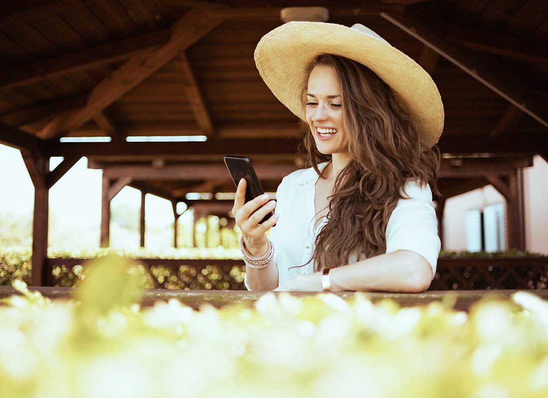 Blog - Closeup Portrait of a Smiling Young Woman Wearing a Straw Hat Standing Outside on a Sunny Day Under a Covered Patio While Using a Phone