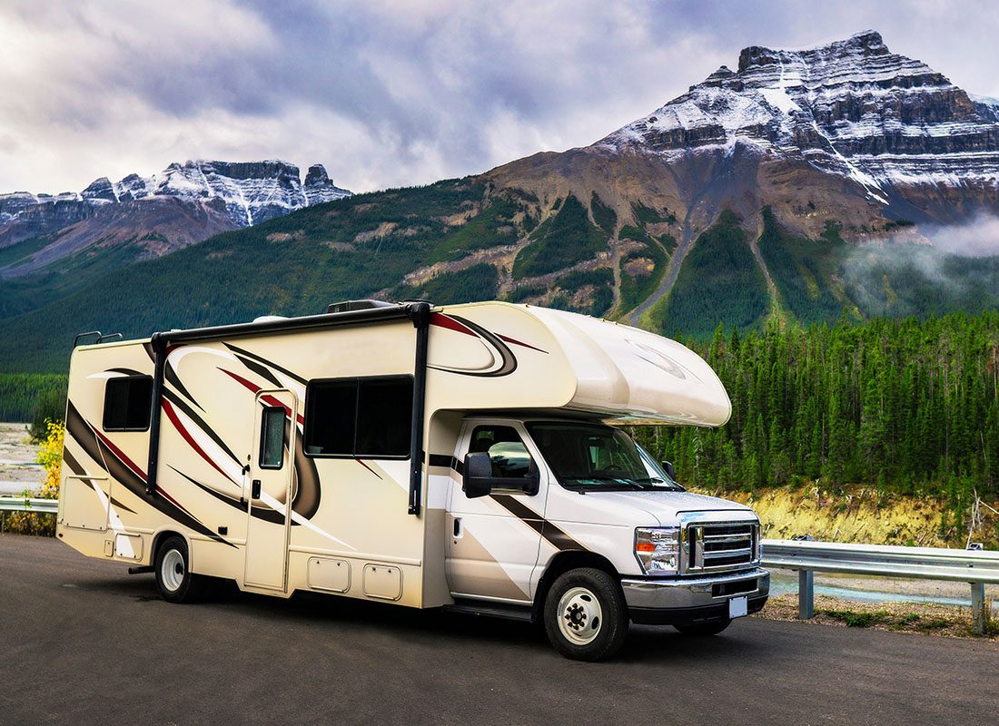 RV Insurance - View of a Small Modern RV Parked Next to a Scenic Road with Evergreen Trees and Snow Capped Mountains in the Background